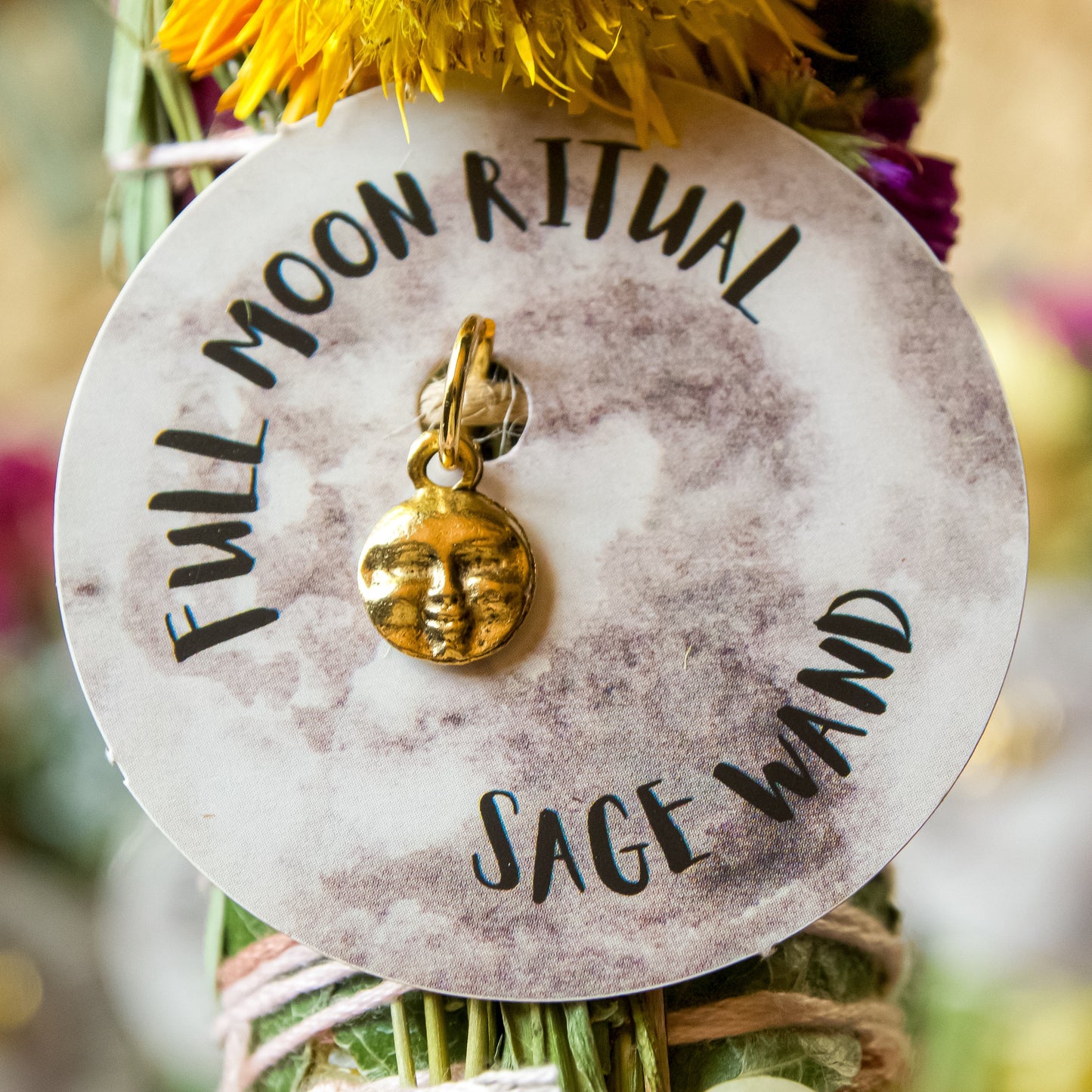 Full Moon Herbal Torch, a hand-wrapped sage wand with ethically sourced white sage, beeswax, local herbs, flowers, and garden greens, with a gold pendant attached.