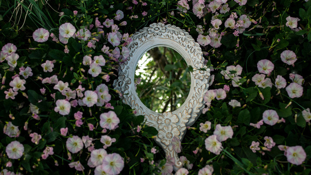 A decorative round mirror with a detailed white frame, reflecting a lush backdrop of light pink morning glory flowers.