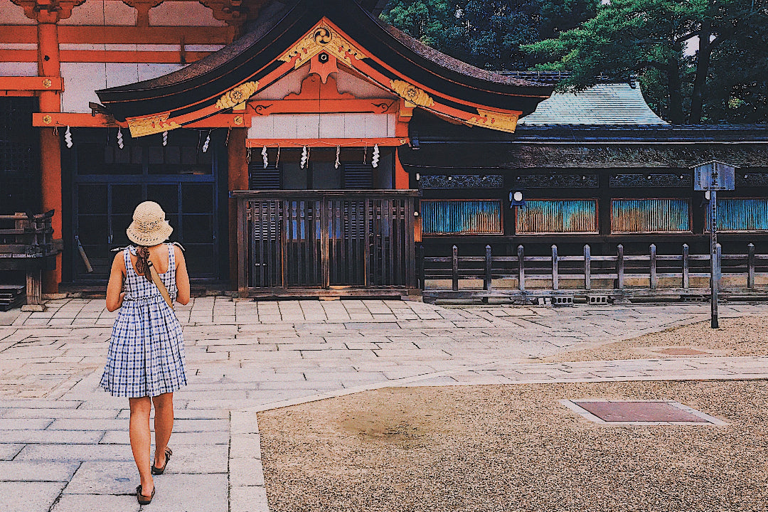 Back view of a young woman in a summer babydoll dress and straw hat, walking towards a Kyoto shrine, symbolizing her journey into awakening intuition and body wisdom.