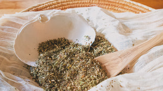 An open cheesecloth displaying a blend of ground dried herbs and flowers, accompanied by a wooden spoon and seashell, evoking the peaceful and soothing experience of tea baths and the practicality of using cheesecloth.