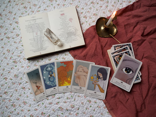Tarot spread with cards laid out on a floral quilt, accompanied by an open book about Venus, Goddess of Love, a candle, and a clear quartz crystal, with the caption 'I love having the Divine on speed dial