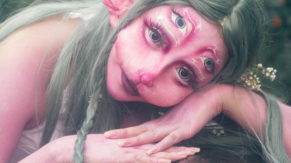 A whimsical fairy-like creature with pink skin, several eyes, and long, green hair, resting its head on its hands