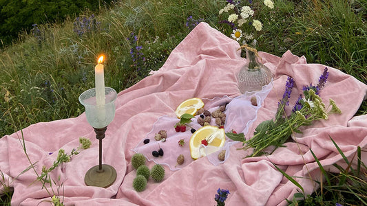 A serene Flower Moon ritual setup in a meadow at dusk, featuring a lit candle on a pink blanket, surrounded by wildflowers, lemons, and berries, creating a peaceful and enchanting atmosphere.