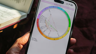 Close-up of a smartphone screen showing the astrological chart for the April 27, 2025 New Moon in Taurus, held in hand with an open book in the background.