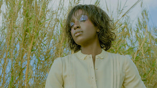 A serene woman with soft makeup and closed eyes, standing amidst tall, wild grasses under a clear sky. This image evokes a sense of calm introspection and connection to nature, representing the reflective energy of the Sturgeon Moon in Aquarius.