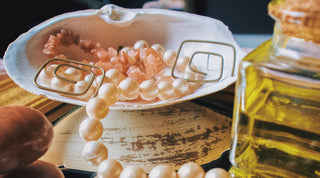 A close-up of pearls and gold earrings nestled within a seashell, surrounded by the soothing scent of essential oil fragrance in a glass bottle. The gleaming jewels reflect on the mirror, creating an aura of sophistication and charm.