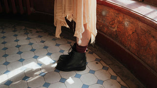 A person wearing a flowing, worn dress and lace-up boots, standing in a vintage tiled room, reflecting an earthy and grounded aesthetic.