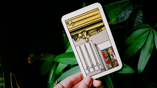 An image of a hand holding the four of swords raider waite tarot card in it's reversed position against a dark background with plants