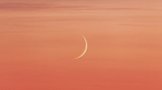 Photo of a peach sherbet-colored sky with a new moon visible in the center of the frame. The image captures the mystical and dreamy energy of the New Moon in Aries Hybrid Solar Eclipse.