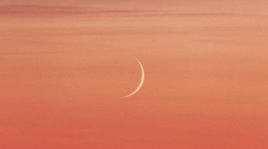 Photo of a peach sherbet-colored sky with a new moon visible in the center of the frame. The image captures the mystical and dreamy energy of the New Moon in Aries Hybrid Solar Eclipse.