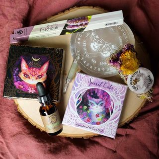 Flat lay of tarot decks, incense, a crystal pendulum, ritual oil, and a sage wand arranged on a wooden slab.